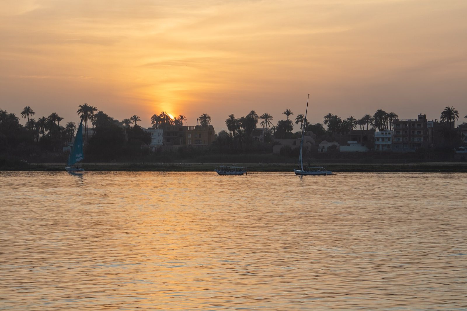 Sunset over the Nile, with a felucca sailing boat on the water and palm trees on the far bank